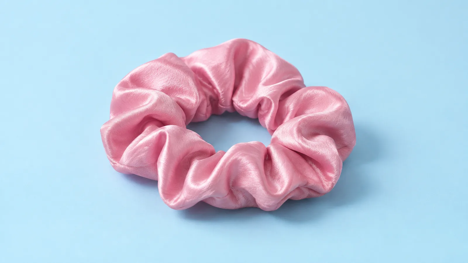 A pink satin scrunchie rests in a loose ring on a pale blue surface, with soft folds and a clearly visible central opening.