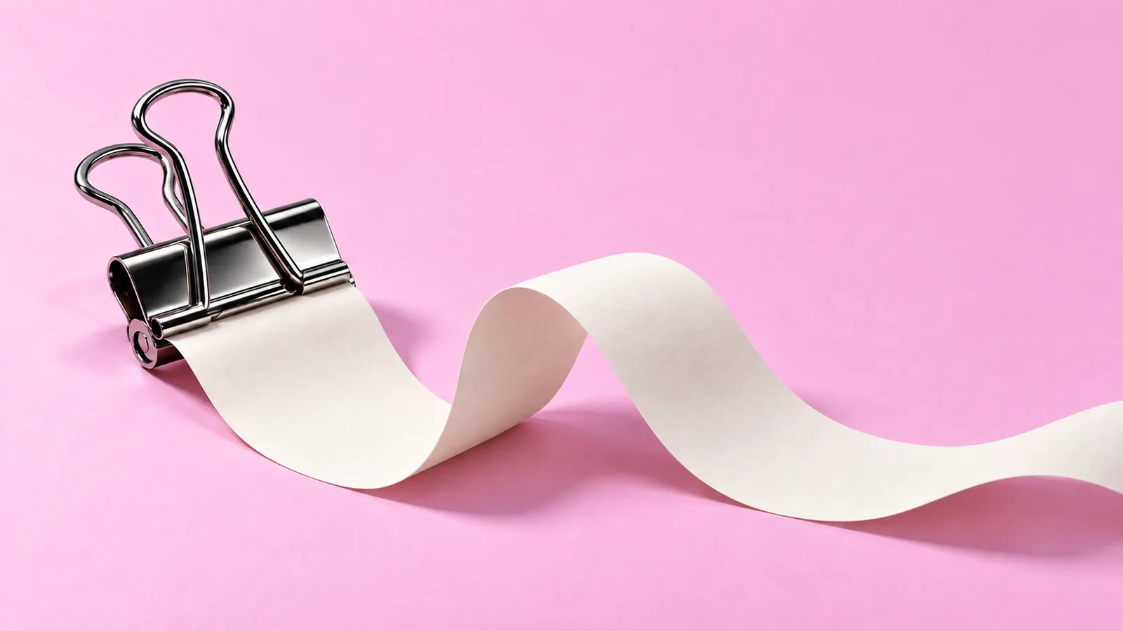 A chrome binder clip holds a continuous cream paper strip that rises in smooth folds and continues beyond the right edge on a pink background.