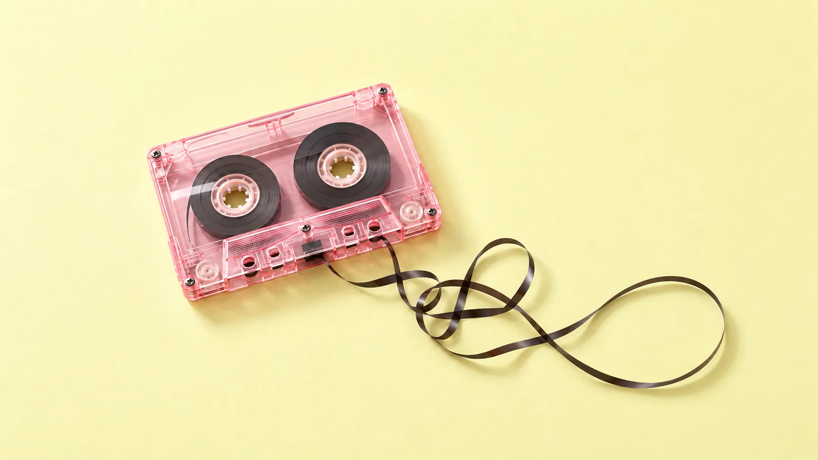A translucent pink cassette rests on a pale lemon-yellow surface, with a thin dark tape unwinding into loose loops beside it.